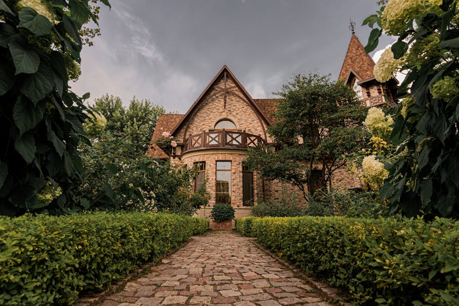 The image shows a large, detached brick house with a steeply pitched roof, timber-framed balcony, and decorative turret, located in a lush garden setting. A cobblestone pathway with a well-maintained hedge on each side leads directly to the front door of the house. Surrounding the pathway are dense green foliage, flowering bushes, and mature trees, including a tree partially blocking the balcony. The sky above is overcast with dark clouds, indicating potential rain. Inside the property, in the foreground, a professional removals team from Man with Van Manor House is actively engaged in a home relocation process, loading cardboard boxes, wrapped furniture, and packing materials into a large van parked outside. The scene captures the logistics of packing, furniture transport, and the loading process during a house move in Manor House, with the garden providing a scenic backdrop to the working environment.