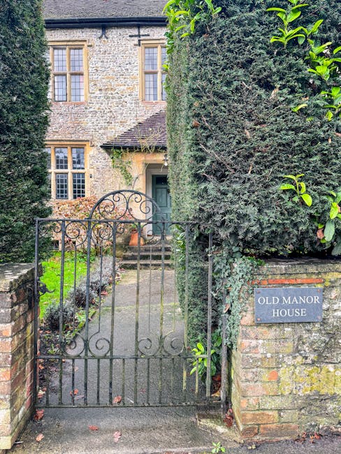 A metal pedestrian gate with decorative scrollwork at the entrance to an old manor house is partially open, revealing a concrete pathway leading to the front door. The gate is mounted between two bric