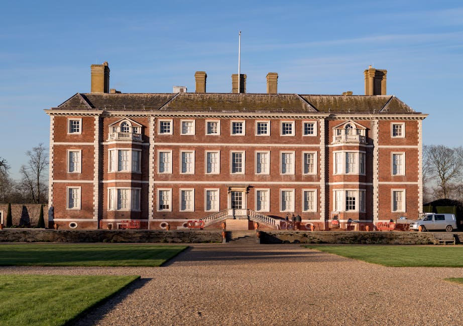 A large, historic brick mansion with symmetrical three-story design, featuring numerous rectangular sash windows, some with decorative white trim, and two small bay windows on either side of centrally located entrance doors. The building has a pitched roof with multiple chimney stacks, and the exterior includes stone detailing along the roofline and around the windows. In front of the mansion, a gravel pathway leads up to a stone staircase with metal handrails, situated in a well-maintained lawn with trimmed grass and a low stone wall. A white van is parked on the driveway to the right side of the building, and a few people are visible near the entrance, engaged in activities related to house removals. The scene is set during daylight with a clear blue sky overhead, and trees are visible in the background, creating a stately environment associated with house removals and relocation services.