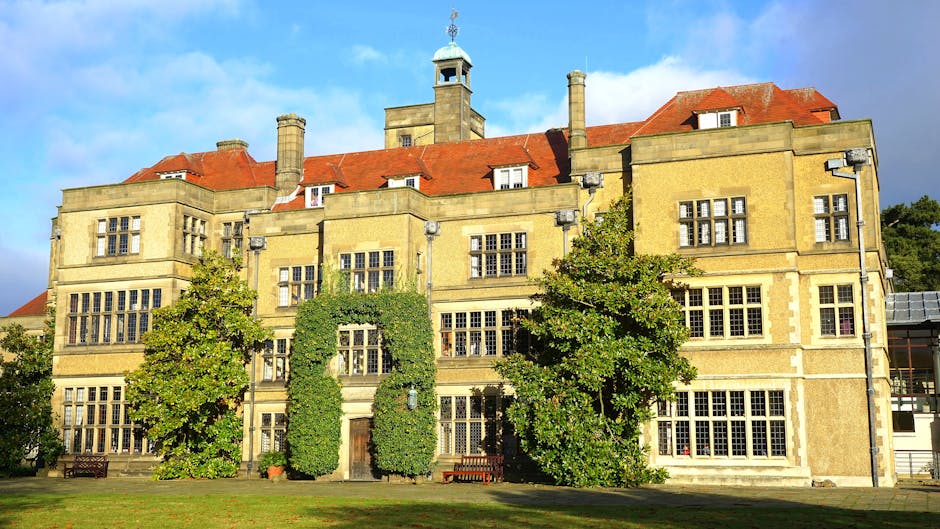 Photograph of a historic, multi-story stone building with large, rectangular windows, some with decorative grid patterns, set against a partly cloudy sky. The building features a red-tiled roof with multiple chimneys, dormer windows, and a small clock tower on top. In front of the building, there are several mature trees with lush green foliage, partially obscuring the ground-floor windows, and two wooden benches visible on either side near the entrance. The scene is set on a well-maintained grassy area, and the image is taken during daylight, highlighting the building’s architectural details. This setting depicts a location suitable for a house removal or relocation service, illustrating the type of historic or residential property handled by companies like Man with Van Manor House during furniture transport and home moves, within a professional and neutral context.