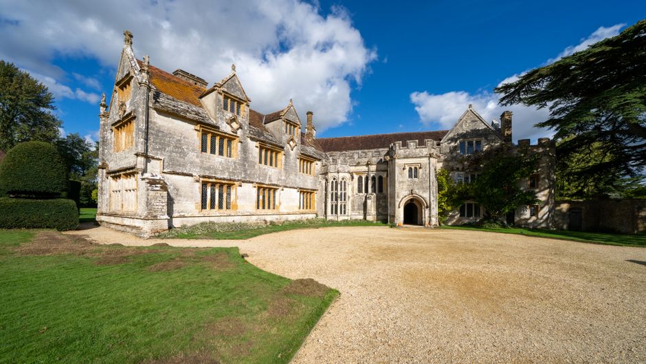 A large historic manor house constructed from grey stone with ornate architectural details, including multiple gabled roofs, stone mullioned windows, and decorative battlements. The building is surrou