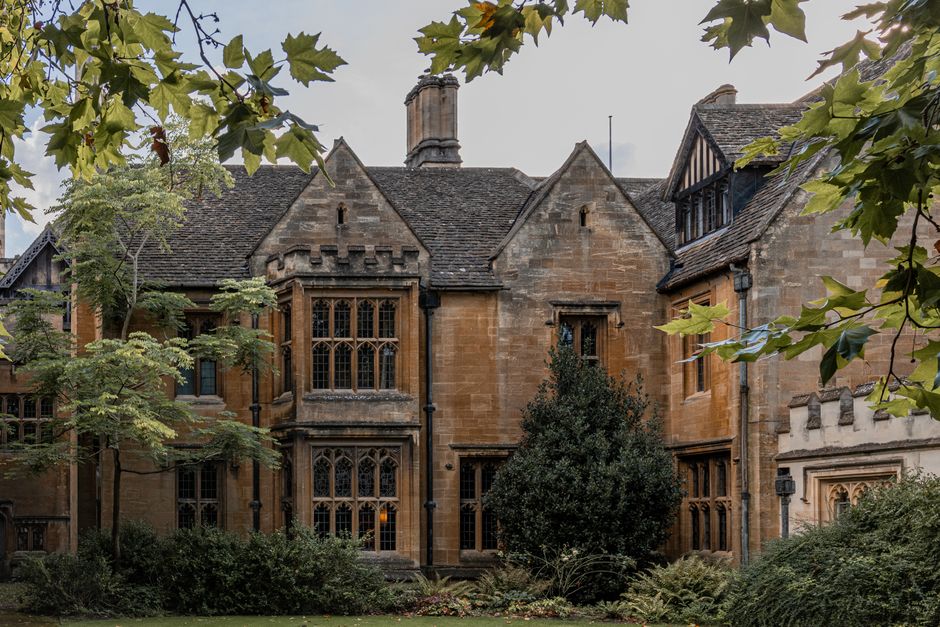 A large historic manor house constructed from grey stone with ornate architectural details, including multiple gabled roofs, stone mullioned windows, and decorative battlements. The building is surrou