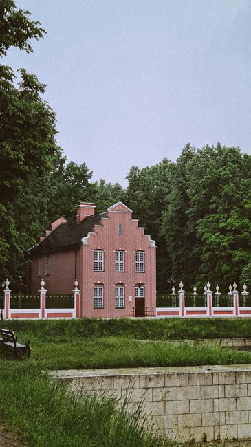 A large, historic stone mansion with multiple levels and numerous tall, rectangular windows, some with arched tops, situated on a grassy area under a cloudy sky. The building features a sloped roof wi