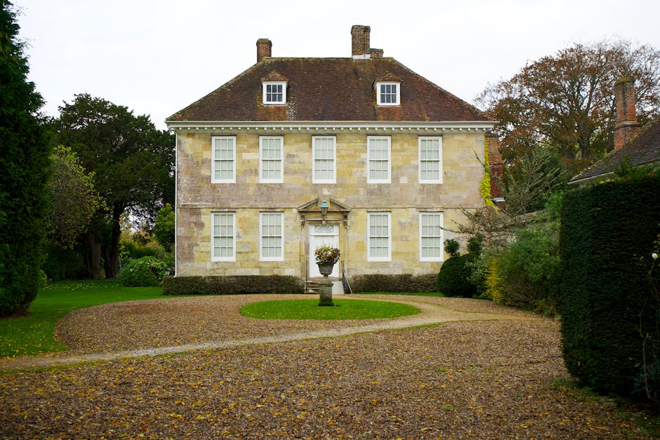 A large Georgian-style house with a symmetrical facade featuring a central doorway and multiple sash windows on each floor, situated on a well-maintained property with a gravel driveway leading to the front entrance. The house has a pitched roof with two chimneys and is constructed from light-colored stone or brick. In front of the house, there is a decorative stone fountain with a planter on top, positioned on a grassy circular patch surrounded by a gravel path. The surrounding garden includes trimmed hedges, mature trees, and lush greenery, with some leafless trees indicating an autumn or early winter season. The lighting is natural, suggesting an overcast day, and the scene captures a peaceful residential setting. This image relates to house removals and furniture transport services offered by Man with Van Manor House, illustrating the typical environment involved during home relocation and packing and moving processes.
