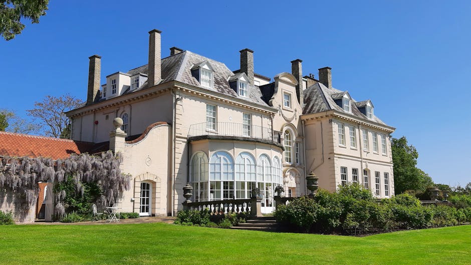 A large, multi-story Victorian-style house with a cream-colored facade, multiple chimneys, and a variety of tall, arched, and rectangular windows. The house features a rounded bay window on the ground floor, surrounded by a low balustrade, and a decorative facade above the entrance. The property is situated on a well-maintained lawn with lush green grass, shrubbery, and flowering plants, including a flowering tree with drooping lavender blossoms on the left side. The sky is clear and blue, indicating a bright, sunny day. The setting appears residential, with no visible vehicles or moving equipment in the immediate vicinity. Man with Van Manor House, specializing in house removals, might be involved in a home relocation process at this property, which could include furniture transport, packing, and loading operations.