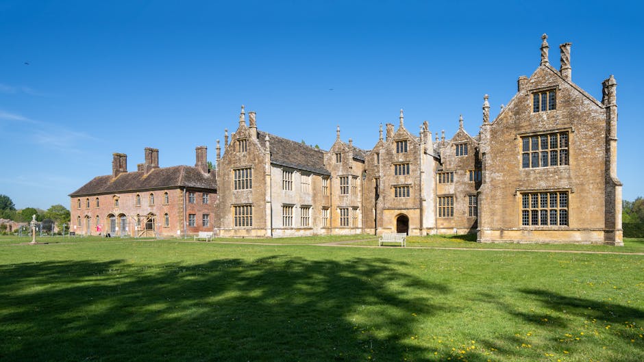 A large historic mansion with a weathered beige facade, multiple decorative gables, and numerous windows set in white frames, situated in a landscaped garden with lush green trees and shrubs. The buil