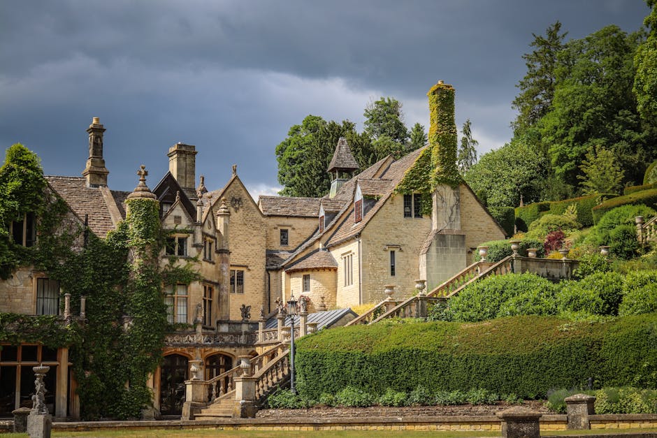 The image shows a historic stone house with multiple chimneys, steeply pitched roofs, and a variety of ornate architectural features, situated on a lush, landscaped hillside. The foreground includes a paved area with stone steps and railings leading from the house to the garden, which is filled with neatly trimmed hedges, vibrant flowering shrubs, and tall trees. The staircase appears to be used for access during a home relocation or moving process, with some areas possibly prepared for furniture transport or packing activities, indicative of ongoing or upcoming house removals. The background features a dark, cloudy sky contrasting with the bright, well-maintained greenery surrounding the property. Man with Van Manor House, a professional removals company, may be involved in the logistics of moving, packing, or furniture transport connected to this residence, as part of a house relocation in N4.