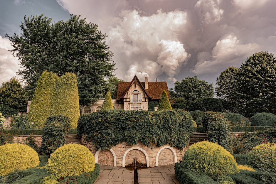 A large, historic building with Victorian architectural features, including decorative cornices, multiple tall sash windows, and a steep, tiled roof with several dormer windows. The building is painted white and surrounded by a low stone wall with a metal railing on top, set within a landscaped area with trees and shrubs. In the foreground, on the pavement or driveway, there are cardboard boxes of various sizes, some sealed with packing tape, arranged near the building's entrance. Additionally, a flatbed trolley with a wooden platform and metal handles is positioned close by, supporting several cardboard boxes for home relocation. A man wearing casual clothing is visible, lifting or carrying a cardboard box toward the building’s entrance, indicating active packing or furniture transport. Nearby, a large moving van with visible side door open, parked on the street, is partially seen, with the vehicle’s rear loading area accessible for furniture transport. The overall scene captures a property being prepared for a house move, with the environment well-lit under natural daylight, emphasizing the process of packing, loading, and logistical preparation for a house removal, related to the services of Man with Van Manor House.