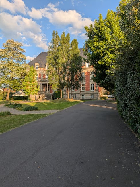 A wide asphalt driveway leading up to a large, historic brick building with multiple stories and architectural details, situated behind a well-maintained lawn and several tall, leafy trees. The sky above is mostly clear with some scattered clouds, and sunlight illuminates the scene, casting shadows from the trees onto the driveway. The building is partially obscured by trees on both sides, and the image appears to be taken during daylight hours. The setting suggests a peaceful, residential area suitable for home relocation or furniture transport, with a clear path for vehicle access to facilitate moving and packing processes, supported by Man with Van Manor House’s professional removals services as referenced on the page about road closure and parking permits.