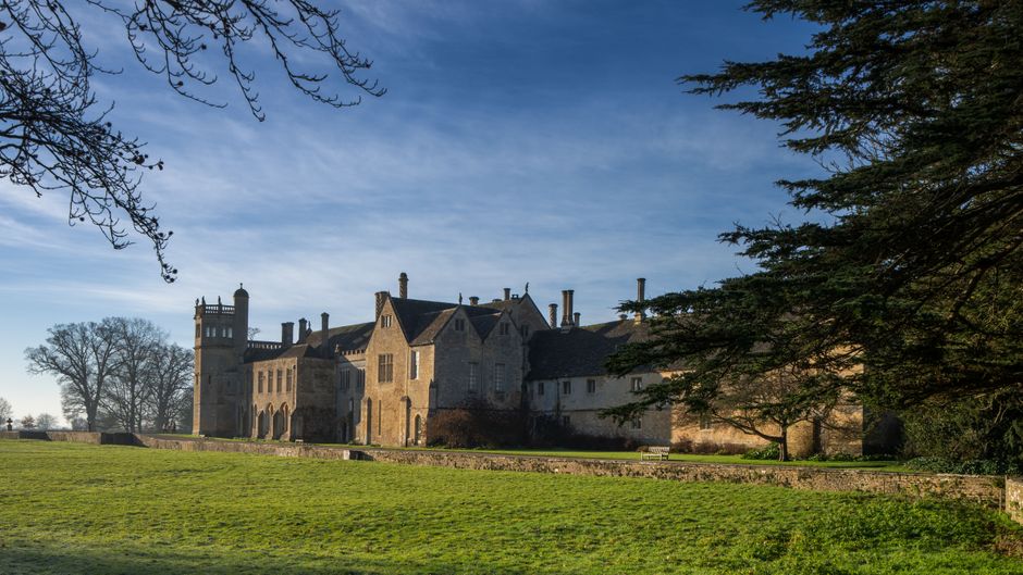 A large historic manor house with stone walls, multiple towers, and chimneys situated in a spacious, well-maintained lawn. The building is partially shaded by large trees with green foliage, and a clear blue sky with wispy clouds is visible overhead. The scene depicts a peaceful and traditional setting, typical of a heritage property that may require professional house removal or moving services. In the foreground, there are no visible vehicles or furniture, but the surrounding area suggests preparations for furniture transport or home relocation. The manor house's exterior shows windows and architectural details consistent with a period estate, with the environment lit by natural daylight. This scene aligns with the context of planning a move or loadings, such as loading furniture into a van outside the property, which is relevant to [COMPANY_NAME]’s removals services on the page about road closure and parking permits for Manor House.