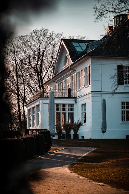 The image shows a large, white, multi-story house with a dark, pitched roof and several large windows, situated amidst leafless trees and a landscaped lawn. In the foreground, a paved driveway curves around the property, with a tall, grey bollard and neatly trimmed hedges lining the edge. The house features a porch area with glass doors and potted plants, possibly indicating an ongoing home relocation or furniture transport process. The lighting suggests early evening or late afternoon, casting shadows across the building and surrounding landscape. The scene reflects the setting for house removals, with a focus on the property's exterior and the surrounding environment, aligning with professional removals services like those provided by Man with Van Manor House.